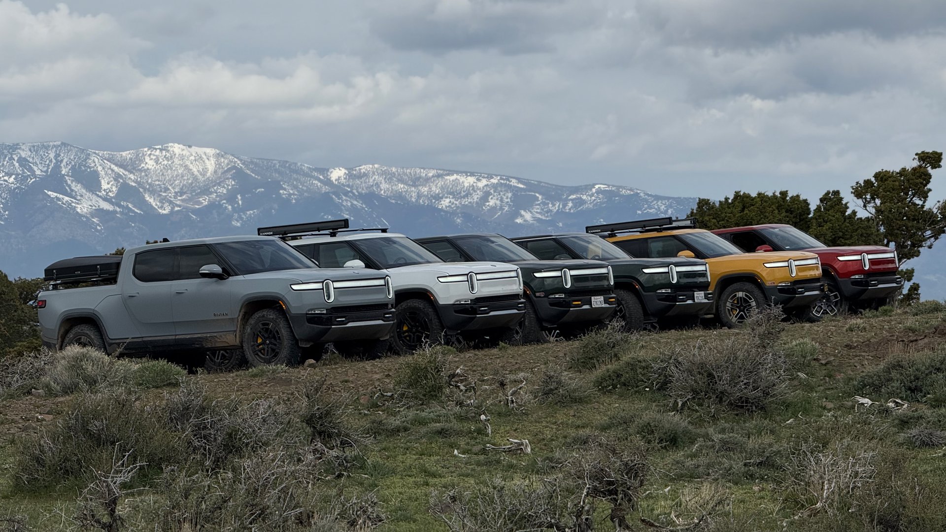 A group of Rivian trucks on a rocky trail below snow-covered mountains